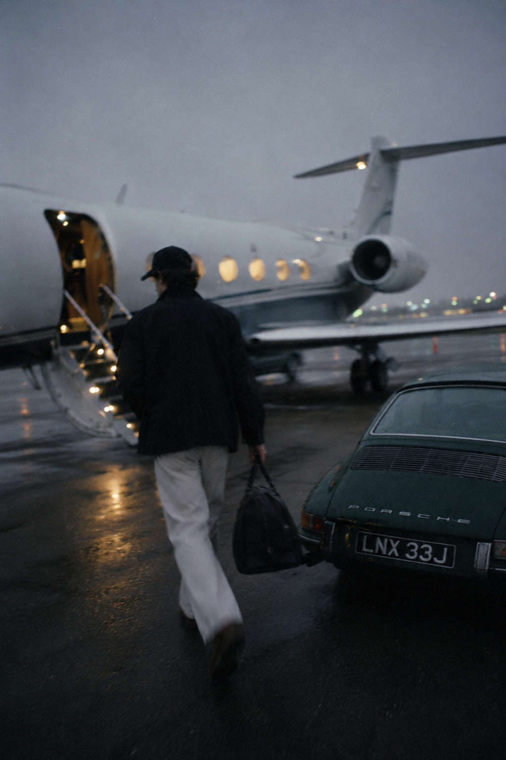 Man in a black cap walking toward a private jet beside a vintage Porsche on a rain-slicked tarmac at dusk